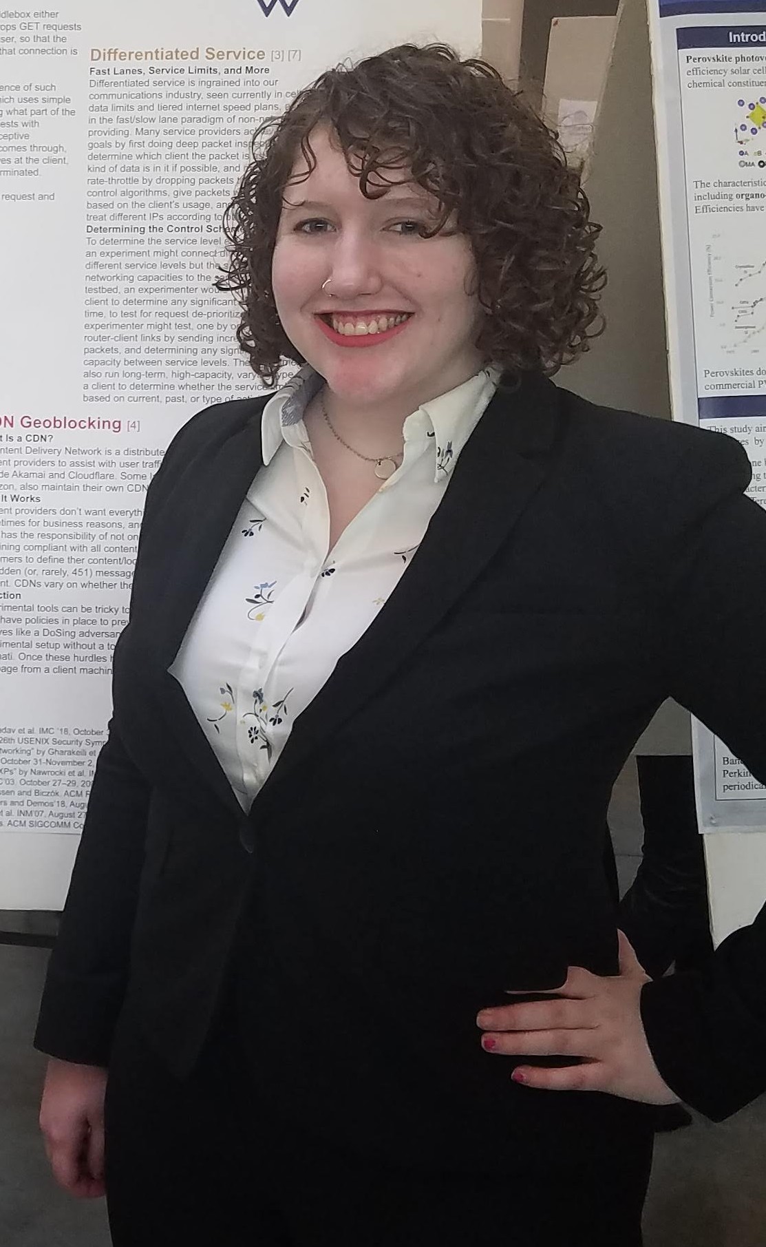 Allison smiles at the camera with her hand on her hip, standing in front of two research posters. She is wearing a dark suit and a white collared blouse sparsely patterned with blue flowers.