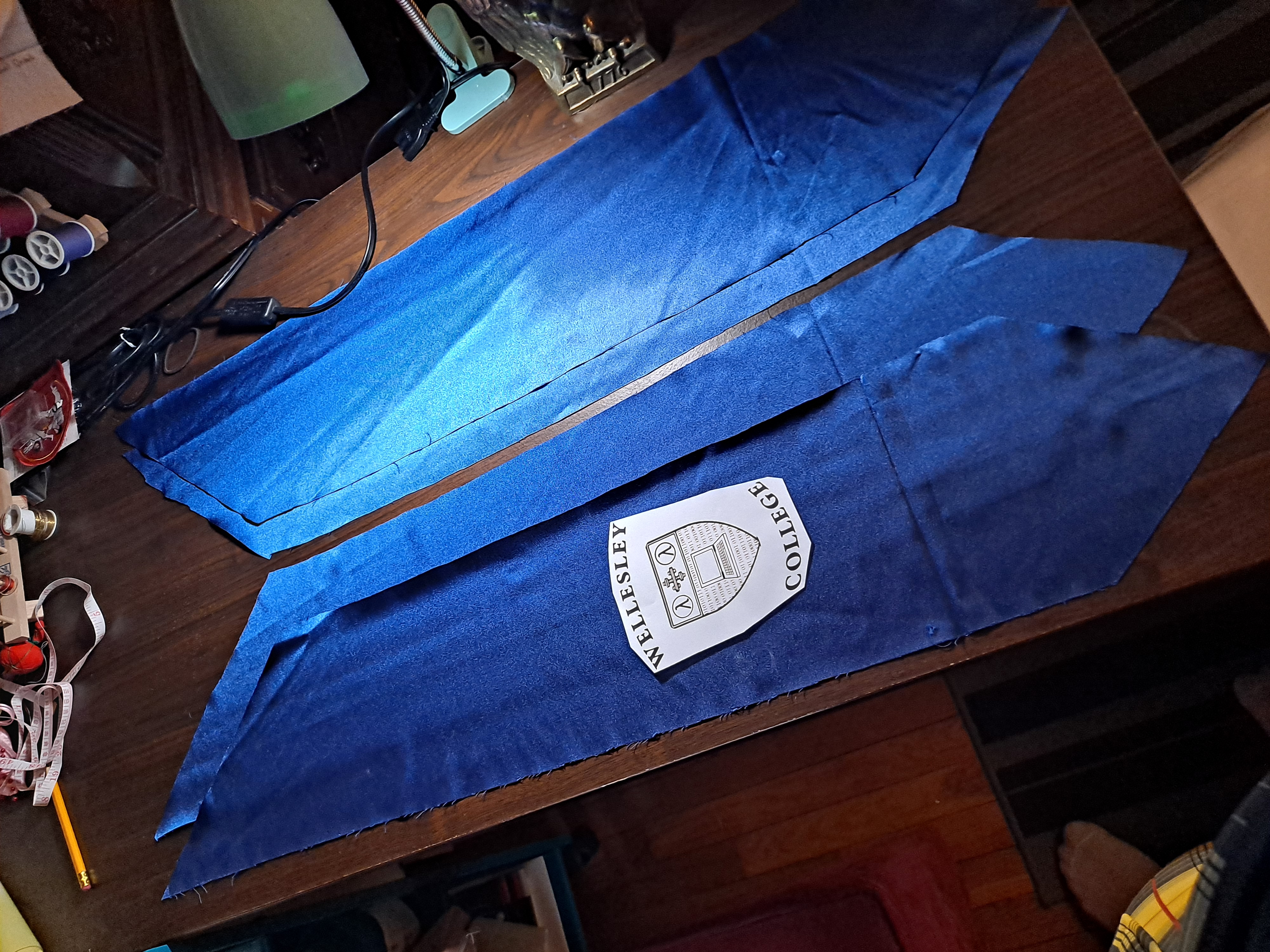 several pieces of blue satin laying on a sewing table, with a paper cutout of the Wellesley CS logo placed on top of one